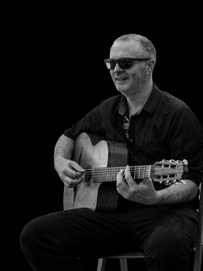 Josh Luke Bray seated with a gypsy jazz guitar, in black and white